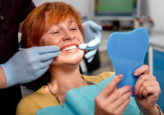 Woman with red hair in dental chair smiling at reflection in mirror with dentist touching corners of her mouth